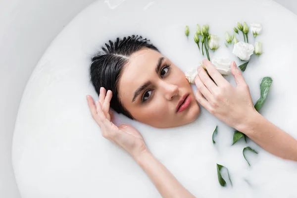 Jovem mulher tocando o cabelo enquanto toma banho no leite com flores eustoma — Fotografia de Stock