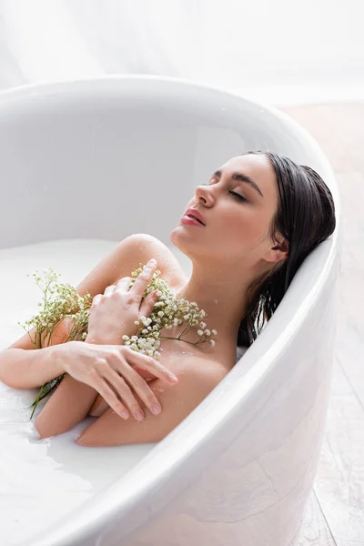 Pretty woman with closed eyes and white, tiny flowers sitting in bathtub with milk — Stock Photo