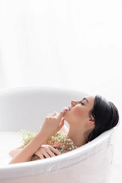 Mujer joven tocando la barbilla y sosteniendo flores de gypsophila mientras se baña en la leche - foto de stock