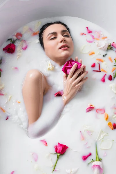 Sensual mujer disfrutando de baño floral con leche mientras sostiene rosa - foto de stock