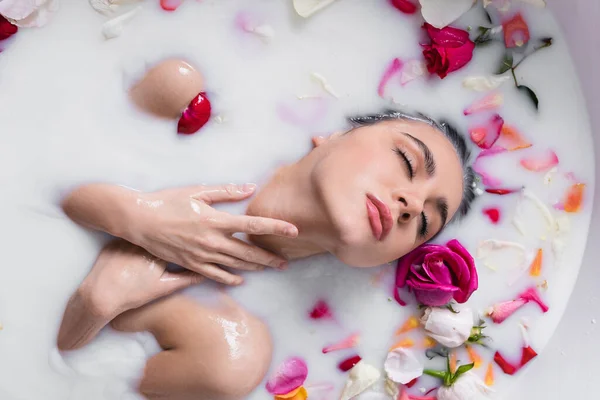 Verführerische Frau, die beim Milchbad mit Rosenblüten den Hals berührt — Stockfoto