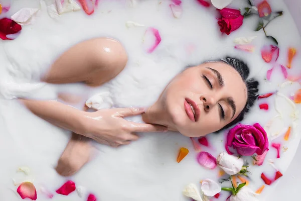 Sensual woman touching neck while taking milk bath with rose petals — Stock Photo