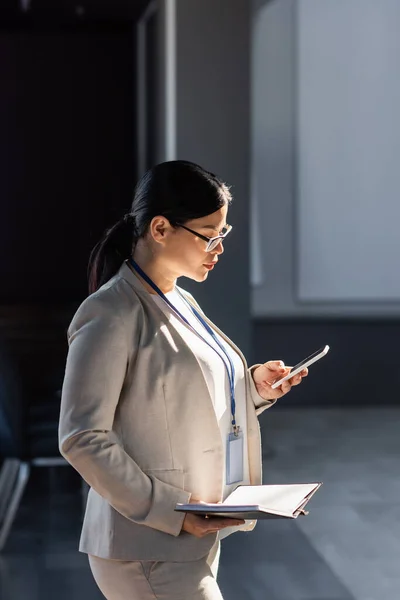 Asian businesswoman in suit using smartphone and holding notebook in office — Stock Photo