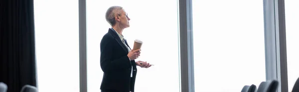 Вид збоку бізнес-леді з паперовою чашкою та мобільним телефоном, що стоїть біля вікна, банер — стокове фото