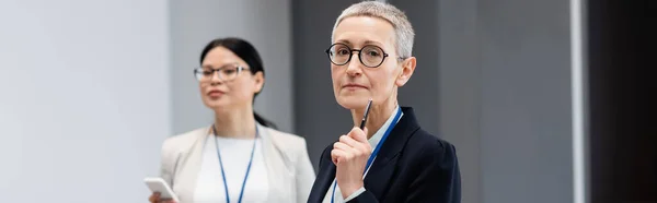 Mature businesswoman with pen standing near blurred asian colleague, banner — Stock Photo