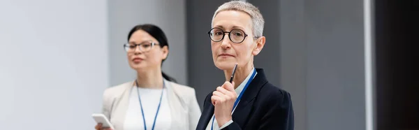 Reife Geschäftsfrau mit Stift steht in der Nähe verschwommenen asiatischen Kollegen, Banner — Stockfoto