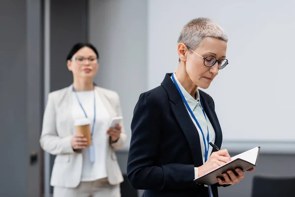 Reife Geschäftsfrau schreibt auf Notizbuch neben asiatischem Kollegen auf verschwommenem Hintergrund — Stockfoto