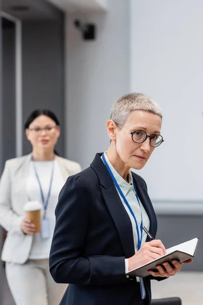 Geschäftsfrau schreibt auf Notizbuch neben verschwommenem Kollegen im Büro — Stockfoto