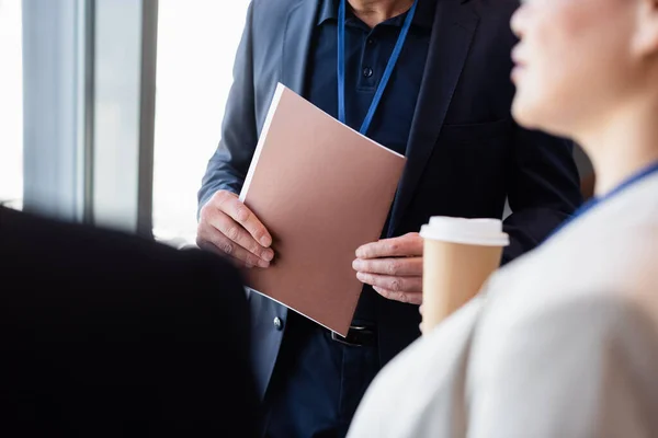 Vista recortada del hombre de negocios sosteniendo carpeta de papel cerca de colega con café para ir en primer plano borrosa - foto de stock