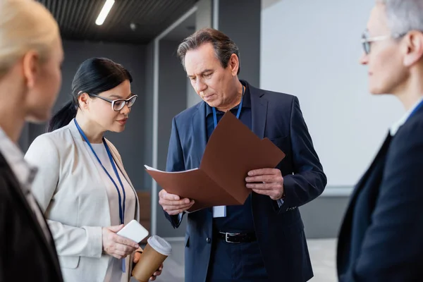 Businessman showing paper folder to asian colleague with coffee to go and smartphone — Stock Photo