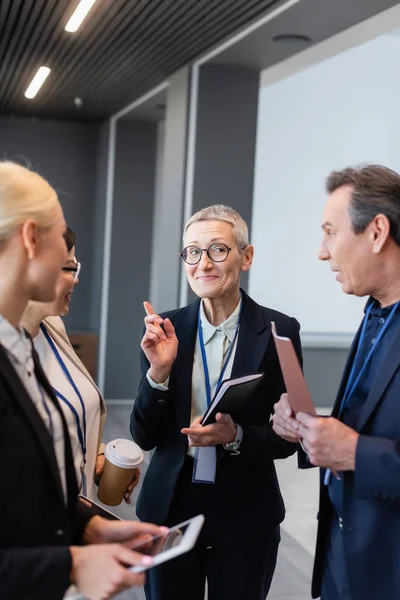 Smiling businesswoman pointing with finger near colleagues holding digital tablet and paper folder — Stock Photo