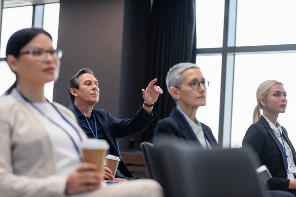 Businessman with coffee to go pointing with finger near colleagues in conference room — Stock Photo