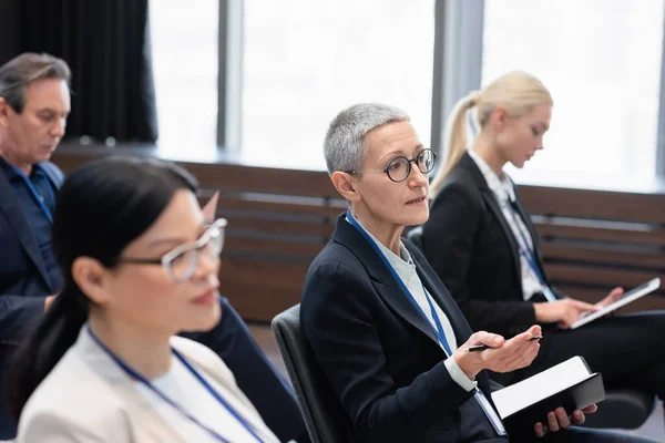 Mature businesswoman with pen and notebook talking during conference — Stock Photo