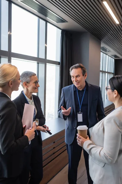 Hombre de negocios sonriente apuntando con la mano cerca de empresarias interracial con smartphone y café para llevar — Stock Photo