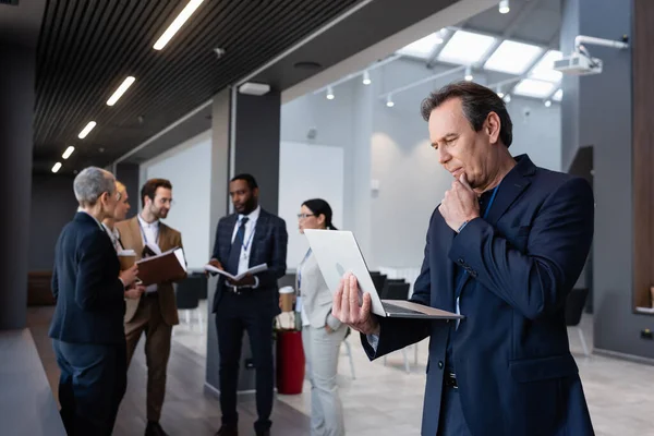 Mature businessman looking at laptop near interracial colleagues on blurred background — Stock Photo