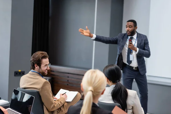 Homme d'affaires souriant avec un cahier regardant un collègue près d'un orateur afro-américain flou dans la salle de conférence — Photo de stock