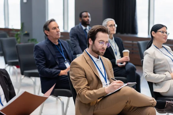 Un homme d'affaires écrit sur un carnet près des hommes d'affaires interracial flous dans la salle de conférence — Photo de stock