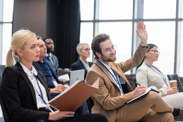 Sonriente hombre de negocios levantando la mano durante el seminario cerca de colegas interracial - foto de stock