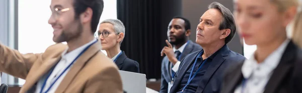 Homme d'affaires d'âge moyen avec ordinateur portable assis près de collègues multiethniques pendant le séminaire, bannière — Photo de stock