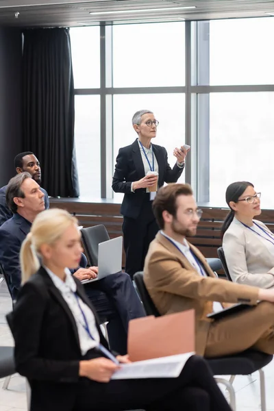 Mature businesswoman with smartphone and coffee to go talking near interracial colleagues during seminar — Stock Photo