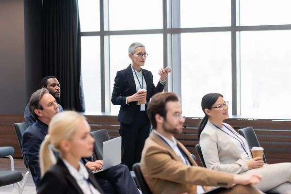Businesswoman with cellphone and takeaway drink talking near multicultural colleagues in conference room — Stock Photo