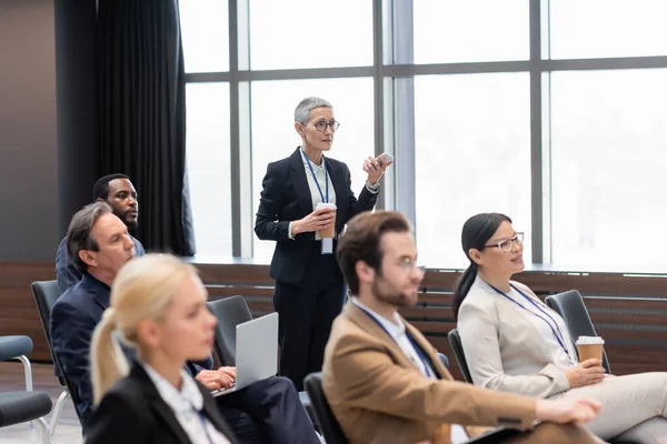 Femme d'affaires avec téléphone portable et boisson à emporter parlant près de collègues multiculturels dans la salle de conférence — Photo de stock