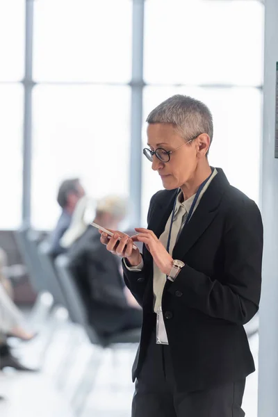 Madura mujer de negocios utilizando teléfono inteligente cerca de la sala de conferencias borrosa - foto de stock