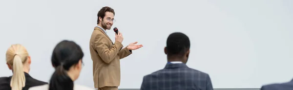 Улыбающийся оратор с микрофоном, указывающий рукой на межрасовых бизнесменов, баннер — стоковое фото