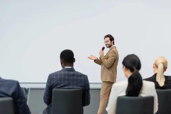 Conférencier souriant parlant et pointant de la main près des gens d'affaires multiethniques flous pendant le séminaire — Photo de stock