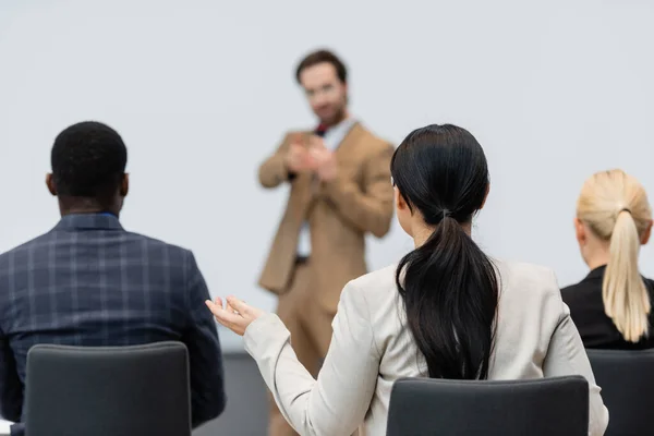 Businesswoman sitting in conference room near interracial colleagues and blurred speaker — Stock Photo