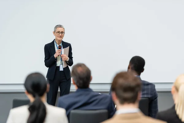 Mujer de negocios sonriente con micrófono y carpeta de papel hablando cerca de gente de negocios borrosa en la sala de conferencias - foto de stock