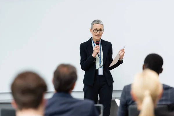 Mature speaker with paper folder talking near blurred business people during meeting — Stock Photo