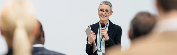 Mature businesswoman with microphone and paper folder talking near business people on blurred foreground, banner — Stock Photo