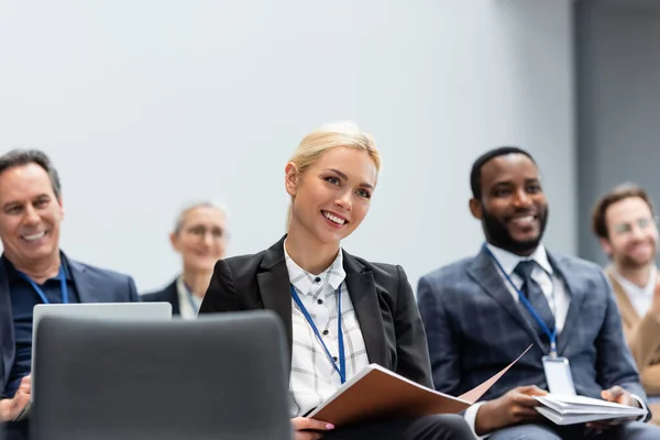 Alegre empresaria con carpeta de papel sentada cerca de colegas multiétnicos durante el seminario — Stock Photo