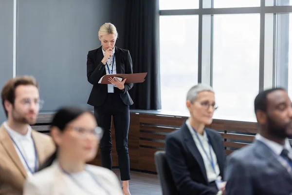 Femme d'affaires avec smartphone et dossier papier debout près des hommes d'affaires interraciaux dans la salle de conférence — Photo de stock