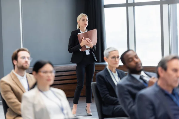 Femme d'affaires avec dossier papier et téléphone portable debout près de collègues multiethniques au premier plan flou — Photo de stock