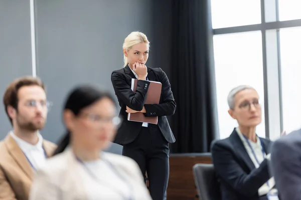 Femme d'affaires concentrée avec dossier papier debout près de collègues flous dans la salle de conférence — Photo de stock