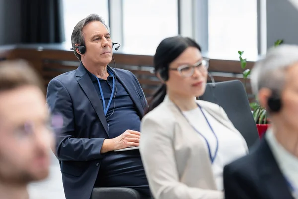 Senior businessman in headset holding laptop near colleagues during conference — Stock Photo