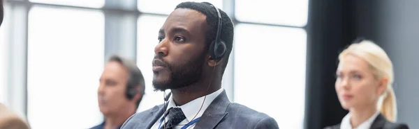 Homme d'affaires afro-américain dans un casque près de collègues flous, bannière — Photo de stock