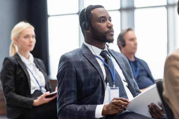 Empresario afroamericano con auriculares sosteniendo papeles cerca de colegas borrosos - foto de stock