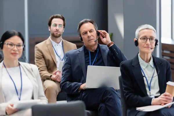 Homem de negócios maduro no fone de ouvido segurando laptop perto de pessoas de negócios inter-raciais na sala de conferências — Fotografia de Stock