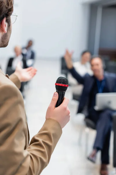 Joven conferenciante con micrófono apuntando al público borroso durante el seminario - foto de stock