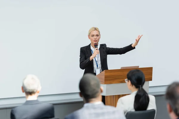 Dozent spricht in Mikrofon und zeigt mit der Hand in die Nähe verschwommener Konferenzteilnehmer — Stockfoto