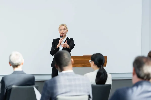 Young lecturer talking in microphone while pointing with finger at business people on blurred foreground — Stock Photo