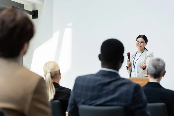 Vue arrière des participants flous près de conférencier asiatique pointant avec la main pendant la conférence d'affaires — Photo de stock