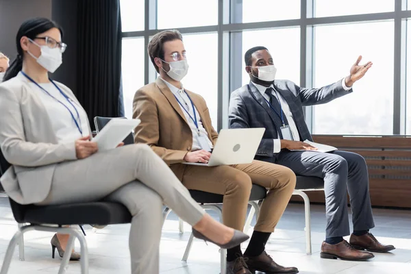 Homme d'affaires afro-américain en masque médical pointant avec la main près de collègues avec des gadgets — Photo de stock
