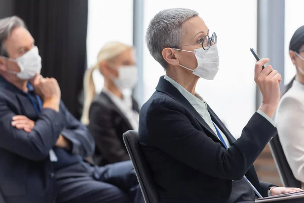 Selektiver Fokus einer reifen Geschäftsfrau in medizinischer Maske, die während eines Seminars einen Stift in der Nähe verschwommener Kollegen hält — Stockfoto