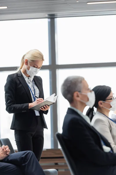 Mujer de negocios en la escritura de la máscara médica en el cuaderno, mientras que de pie en la sala de conferencias - foto de stock