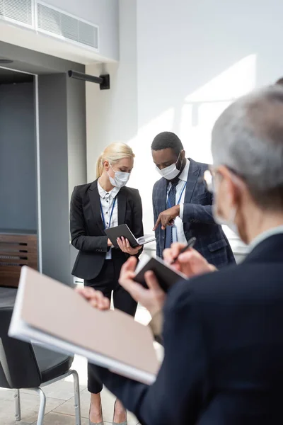 Hombre de negocios afroamericano en máscara médica apuntando a la libreta en manos de un colega, borrosa primer plano - foto de stock