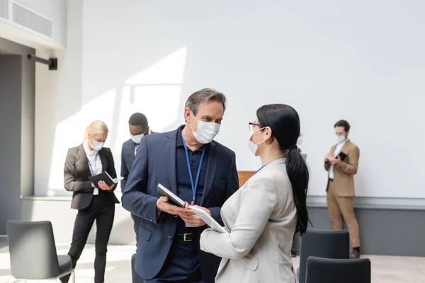 Homem de negócios de meia idade em máscara médica falando com colega asiático durante conferência — Fotografia de Stock
